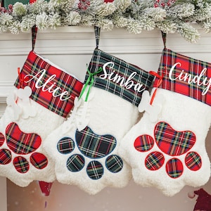 May include: Three Christmas stockings in the shape of a paw, each with a different name: "Alice", "Simba", and "Cindy". The stockings have a red and green plaid pattern and a white fluffy base. A festive holiday decoration.
