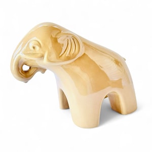 May include: A ceramic elephant figurine in a glossy, light tan color. The elephant has a trunk, ears, and a curved back. The figurine is a decorative item, likely for a shelf or table.