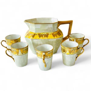 May include: A vintage tea set featuring a pale green pitcher and six matching cups. The set has gold accents on the handles and rims, with a yellow band featuring a grape design around the pitcher and cups. The set is designed for serving tea or coffee.