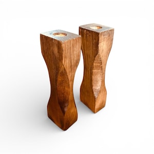 May include: Two wooden candle holders with a square top and base, and a curved, hourglass-like shape. The candle holders are stained a warm brown color, and the tops have a small circular indentation for candles.