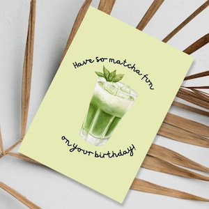 May include: A light green greeting card with the text "Have so matcha fun on your birthday!" features a watercolor illustration of a matcha drink with ice and mint leaves. The card is set against a background of dried palm leaves.
