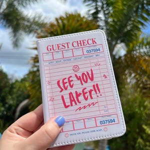 May include: A white guest check book with pink text that reads "GUEST CHECK" and "SEE YOU LATER!!". The check number is 037594. The book is held in a hand with blue nail polish.