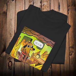 May include: Black t-shirts featuring a cartoon dog in a burning room, with the text "THIS IS FINE" in a speech bubble. The dog is wearing a hat and sitting on a chair, with a coffee cup on a table.