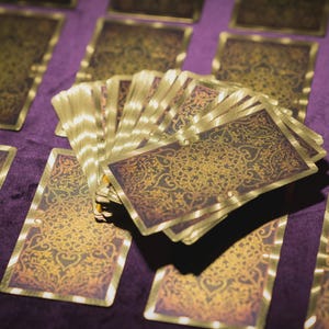 Può includere: Un mazzo di carte dei tarocchi con disegni dorati e marroni ornati, a ventaglio su una superficie di velluto viola. Diverse altre carte sono disposte attorno al mazzo a ventaglio, mostrando l'opera d'arte dettagliata.