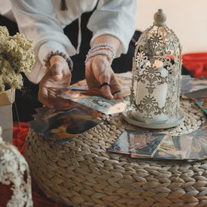 Può includere: Un portacandele decorativo in metallo ornato con una candela accesa all'interno è posto su una superficie intrecciata, circondato da carte dei tarocchi. Fiori secchi in un cono di carta e un secondo portacandele simile sono anch'essi presenti. La scena è ambientata su un tessuto rosso.