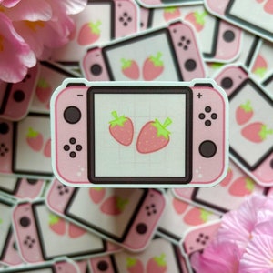 May include: Pink sticker resembling a game console, featuring a screen with two pink strawberries. The sticker is surrounded by other similar stickers and pink flowers, creating a playful and colorful aesthetic. The design is likely aimed at gamers.