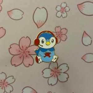 May include: A blue, white, and red penguin pin wearing a headset and holding a game controller. The pin has gold-colored trim. The background is pink with cherry blossom designs.