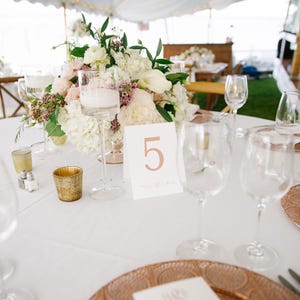 Peut inclure: Une table blanche avec une nappe blanche est dress&eacute;e pour un d&icirc;ner de mariage. Un centre de table compos&eacute; de fleurs blanches et roses est plac&eacute; dans un vase en verre transparent. Le num&eacute;ro de table est "5" et est &eacute;crit en cuivre. Il y a deux assiettes rondes en cuivre sur la table.