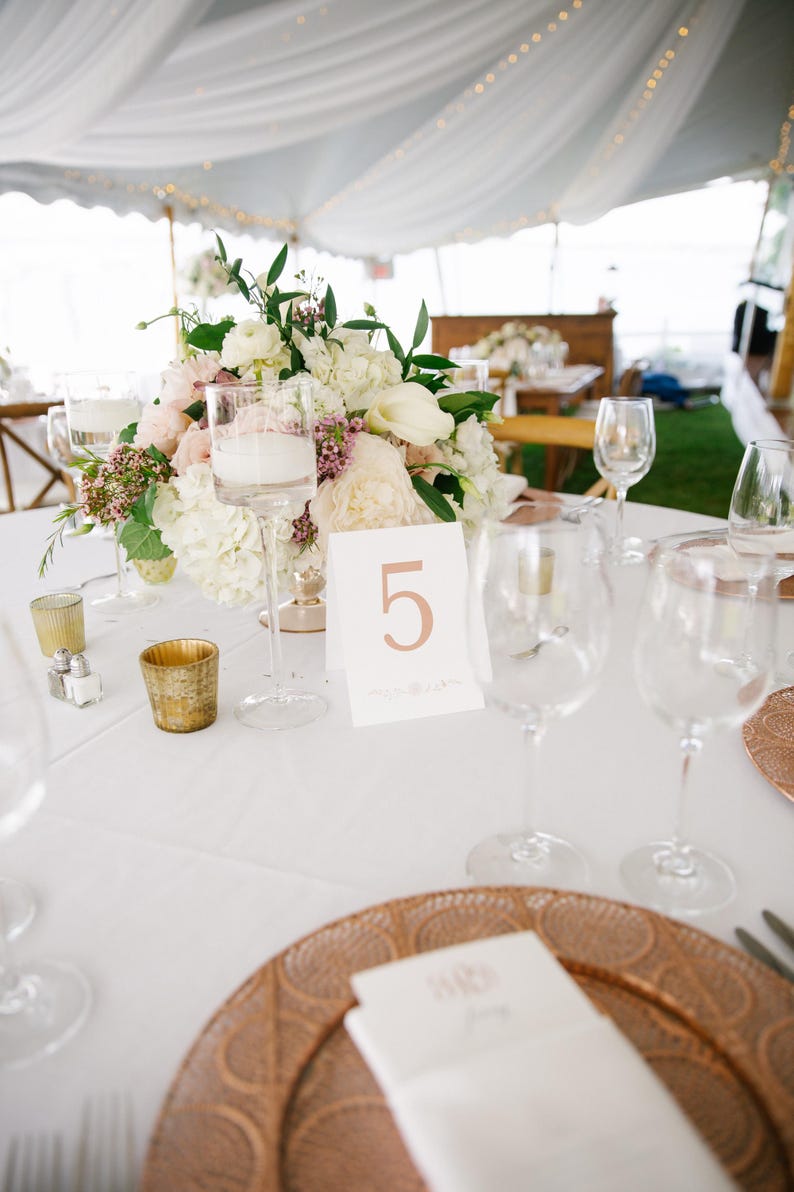 Peut inclure: Une table blanche avec une nappe blanche est dress&eacute;e pour un d&icirc;ner de mariage. Un centre de table compos&eacute; de fleurs blanches et roses est plac&eacute; dans un vase en verre transparent. Le num&eacute;ro de table est "5" et est &eacute;crit en cuivre. Il y a deux assiettes rondes en cuivre sur la table.