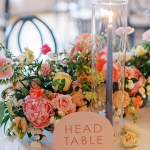 Peut inclure: Un num&eacute;ro de table rose avec le texte "HEAD TABLE" dans une police moderne. Le panneau est plac&eacute; sur une nappe blanche avec une composition florale compos&eacute;e de fleurs roses, p&ecirc;che et jaunes. Un grand cylindre en verre avec une bougie allum&eacute;e se trouve au centre de l'arrangement.