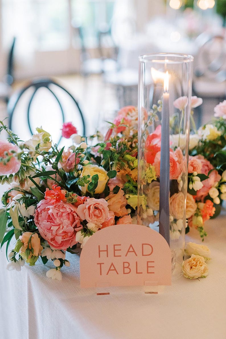 Peut inclure: Un num&eacute;ro de table rose avec le texte "HEAD TABLE" dans une police moderne. Le panneau est plac&eacute; sur une nappe blanche avec une composition florale compos&eacute;e de fleurs roses, p&ecirc;che et jaunes. Un grand cylindre en verre avec une bougie allum&eacute;e se trouve au centre de l'arrangement.
