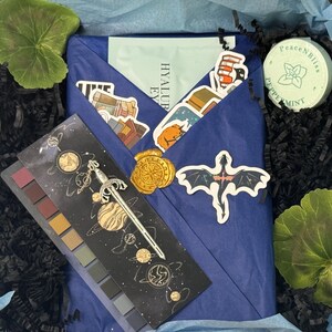 May include: A blue box filled with a variety of items, including a book wrapped in blue paper, a small round container with the text "Peace N Bliss Peppermint", a small palette of eyeshadows with a space-themed design, and a variety of stickers.