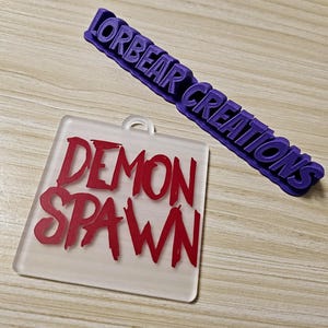 May include: A clear acrylic keychain with the words "DEMON SPAWN" in red lettering. The keychain is next to a purple plastic sign that reads "LORBEAR CREATIONS".