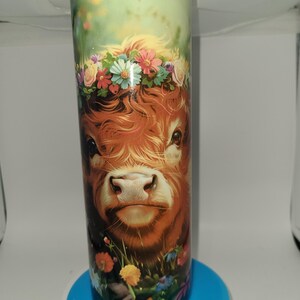 Könnte beinhalten: Ein zylindrischer Becher mit einer detaillierten Illustration einer Highland-Kuh mit Blumenkranz. Die Kuh hat rotbraunes Fell und ist von bunten Blumen umgeben. Der Hintergrund ist ein sanftes Grün, was eine skurrile und ländliche Szene schafft.