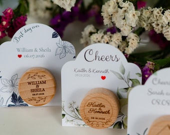 May include: Wooden wedding favours with personalised names and dates. Each favour is a small, round wooden disc with engraved text, paired with a decorative card. The cards feature floral designs and the words "Best day ever" and "Cheers".