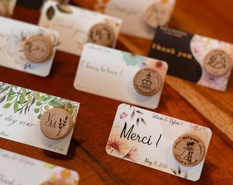 May include: A set of wedding favour cards with floral designs and personalised wooden cork stoppers. The cards feature the text "Merci!" and "May 31, 2026" with the names "Jillian & Dylan".