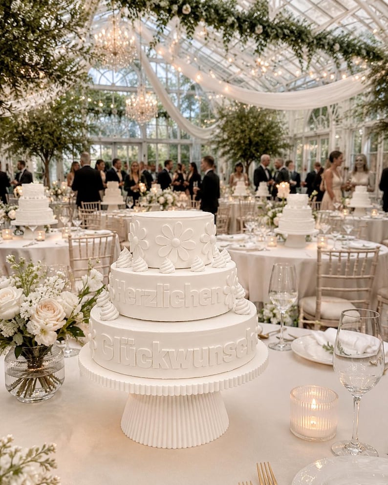 May include: A three-tiered white cake on a fluted pedestal stand. The top tier features embossed daisy flowers, while the middle and bottom tiers display the embossed German text "Herzlichen Gl&uuml;ckwunsch" with small piped frosting accents.