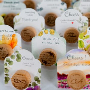 May include: A collection of wooden bottle stoppers with decorative cards. The cards feature floral designs, the words "Thank You" and "Cheers", and names with dates. The stoppers have engraved initials.