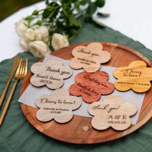 May include: Flower-shaped wooden coasters with custom text, including names and dates. The coasters are displayed on a wooden tray, with a green cloth and gold cutlery in the background. White flowers are also visible.