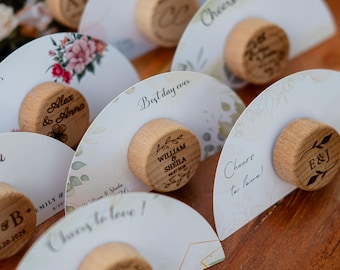 May include: Wooden bottle stoppers with personalised engravings and decorative paper fans. The stoppers feature names, initials, and dates. The fans have floral and botanical designs, with phrases like "Best day ever" and "Cheers to love!"