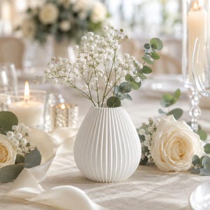 Puede incluir: Un jarr&oacute;n de cer&aacute;mica blanca, acanalado, contiene delicadas flores blancas y hojas verdes de eucalipto. El jarr&oacute;n est&aacute; sobre un mantel crema, con una vela encendida y una rosa blanca en el fondo. La escena evoca elegancia y serenidad.