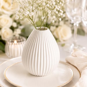 Puede incluir: Un jarr&oacute;n de cer&aacute;mica blanco y acanalado que contiene flores blancas de paniculata. El jarr&oacute;n est&aacute; sobre un plato blanco con borde dorado, parte de una mesa con una vela y flores en el fondo.