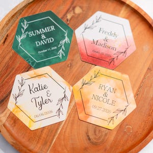 May include: Four hexagonal coasters with watercolor backgrounds in green, red, yellow, and orange, each with names and dates. The coasters are on a wooden tray. The text includes names like Summer & David and dates like October 11, 2026.