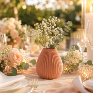 Puede incluir: Un jarr&oacute;n de cer&aacute;mica acanalada de color melocot&oacute;n que contiene flores blancas y vegetaci&oacute;n. El jarr&oacute;n es el punto focal de una mesa, con otros arreglos florales, velas y cristaler&iacute;a en el fondo. La escena est&aacute; ba&ntilde;ada por una suave luz natural.