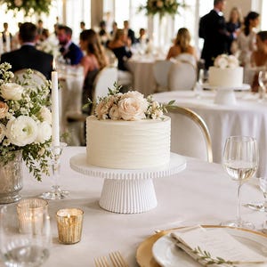 May include: A round white cake with a textured horizontal wave pattern on the sides, topped with a cluster of pale pink roses and greenery, displayed on a white fluted pedestal cake stand on a table set with gold-rimmed plates and candles.