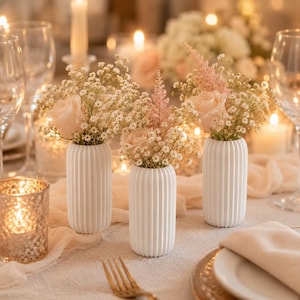 May include: Three white cylindrical vases with vertical ribbed textures, each containing a floral arrangement of light pink roses and small white baby's breath flowers, displayed on a table setting with glassware and soft ambient lighting.