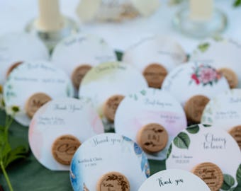 May include: A selection of circular cards with wooden details, possibly for a wedding. The cards display text such as "Thank you" and names, alongside floral or watercolour designs. They are arranged on a green surface, with candles in the background.
