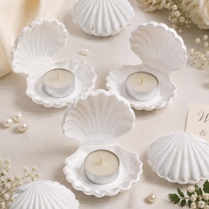 Peut inclure: Une collection de bougeoirs blancs en forme de coquillage, certains ouverts et d'autres fermés, contenant chacun une petite bougie chauffe-plat ronde. L'ensemble est disposé avec des perles blanches et de petites fleurs blanches éparpillées.