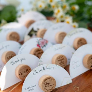 Può includere: Una collezione di biglietti bianchi a forma di semicerchio con tappi per bottiglie in legno, disposti su un vassoio di legno. I biglietti presentano testi come "Thank you" e "Cheers to love!" con nomi e date. Sono visibili anche elementi floreali.