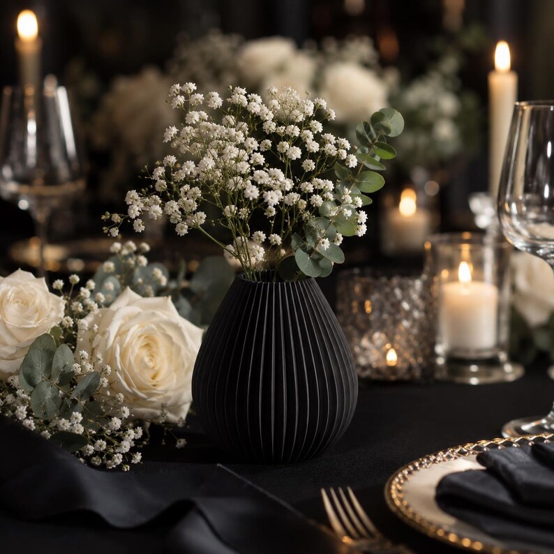 Puede incluir: Un jarr&oacute;n negro con l&iacute;neas verticales que contiene flores blancas y ramitas verdes se encuentra sobre un mantel negro. Rosas blancas y velas en el fondo, con un plato con borde dorado y cubiertos en primer plano. La escena evoca una cena formal.