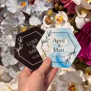 May include: Two hexagonal coasters, one black and one blue, held in front of a floral background. The black coaster has white text that reads "LUKE & SAR" and "10.18.26". The blue coaster reads "April & Matt" and "10/10/2026".