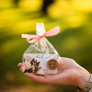 May include: A small, clear plastic bag with a pink ribbon tied around it. The bag contains a white card with a floral design and the text "thank you!" and a small wooden circle with the text "A&E 06.07.23".