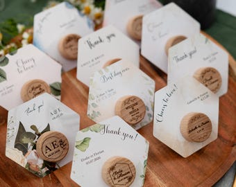 May include: A wooden tray displays several hexagonal cards with wooden round toppers. The cards feature floral designs and text like "Thank you!" and "Cheers to love!" Some toppers have initials and dates.