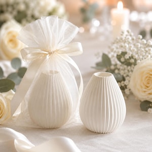 Puede incluir: Dos peque&ntilde;os jarrones blancos acanalados. Uno est&aacute; dentro de una bolsa transparente color crema atada con una cinta a juego. Los jarrones tienen forma de l&aacute;grima y est&aacute;n sobre una superficie blanca, con rosas blancas y velas al fondo.