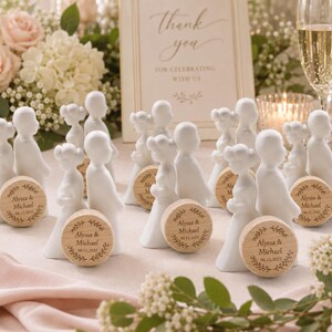 May include: White ceramic figurines of a bride and groom with wooden coasters. The coasters are engraved with names and a date, surrounded by a leaf design. The background includes flowers, champagne glasses, and a sign that says "Thank You".