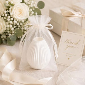 Puede incluir: Un jarr&oacute;n de cer&aacute;mica blanco y acanalado se presenta en una bolsa transparente blanca con cord&oacute;n. Una peque&ntilde;a tarjeta con letras doradas dice "gracias" y una caja de regalo con una cinta est&aacute; en el fondo. Tambi&eacute;n se ve un ramo de rosas blancas y vegetaci&oacute;n.
