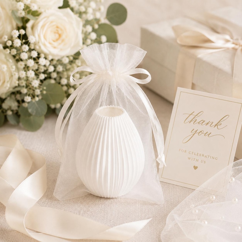 Puede incluir: Un jarr&oacute;n de cer&aacute;mica blanco y acanalado se presenta en una bolsa transparente blanca con cord&oacute;n. Una peque&ntilde;a tarjeta con letras doradas dice "gracias" y una caja de regalo con una cinta est&aacute; en el fondo. Tambi&eacute;n se ve un ramo de rosas blancas y vegetaci&oacute;n.