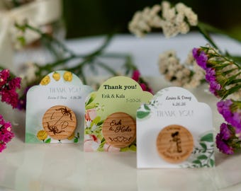 May include: Three wooden wedding favour magnets with floral and botanical designs. Each magnet has a unique design and the words "Thank you" printed on the card. Names and dates are also printed on the cards.