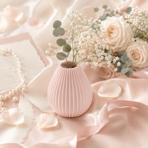 Puede incluir: Un peque&ntilde;o jarr&oacute;n rosa p&aacute;lido, acanalado, contiene flores blancas y follaje. El jarr&oacute;n est&aacute; sobre una tela rosa suave, con p&eacute;talos de rosa dispersos, un collar de perlas y un ramo de rosas crema en el fondo. La escena evoca elegancia.