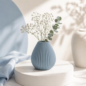 Puede incluir: Un jarr&oacute;n de cer&aacute;mica azul claro y acanalado con flores blancas y hojas de eucalipto verdes. El jarr&oacute;n, con forma de l&aacute;grima, est&aacute; sobre un pedestal blanco. El fondo incluye tela azul suave y un jarr&oacute;n blanco.