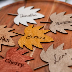 Puede incluir: Tarjetas de lugar con forma de hoja de madera con nombres grabados. Las hojas son de varios colores, incluyendo blanco, naranja y marrón, y están dispuestas sobre una bandeja de madera. Los nombres incluyen Jonathan, Brittany y Cherry.