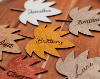 Puede incluir: Tarjetas de lugar con forma de hoja de madera con nombres grabados. Las hojas son de varios colores, incluyendo blanco, naranja y marrón, y están dispuestas sobre una bandeja de madera. Los nombres incluyen Jonathan, Brittany y Cherry.
