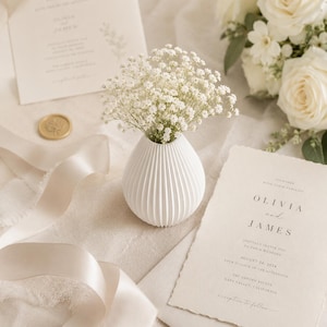Puede incluir: Un jarr&oacute;n de cer&aacute;mica blanco acanalado con peque&ntilde;as flores blancas, invitaciones de boda y un sello de cera dorado. La invitaci&oacute;n dice "Olivia y James" y la fecha "24 de agosto de 2024". Cintas suaves y rosas blancas completan la escena.