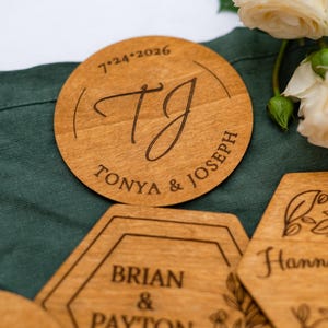 May include: Wooden wedding coasters with engraved names and dates. One coaster is round with "TJ" and "TONYA & JOSEPH" with the date "7.24.2026". Other coasters are hexagonal with names and dates.