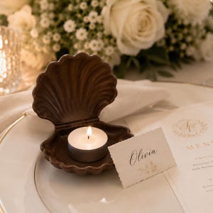 May include: A dark brown, shell-shaped candle holder made of a rigid material, holding a small, round, white tea light candle with a lit flame. The shell is open, revealing the candle inside, and is placed on a white plate next to a place card.