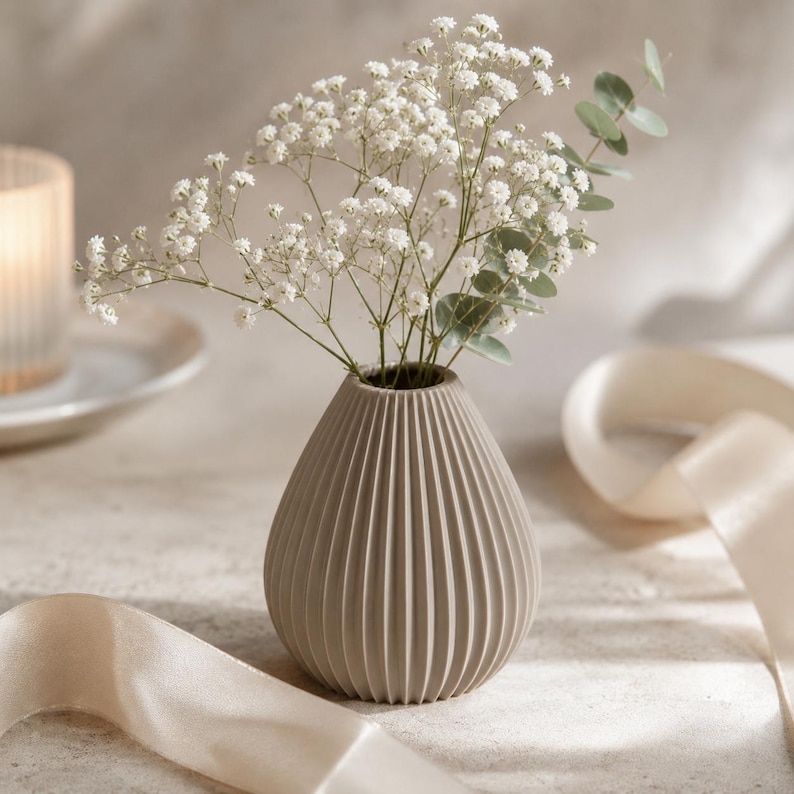 Puede incluir: Un peque&ntilde;o jarr&oacute;n beige y acanalado contiene un ramo de flores blancas y hojas verdes de eucalipto. El jarr&oacute;n tiene forma de l&aacute;grima. Una vela y una cinta est&aacute;n en el fondo.